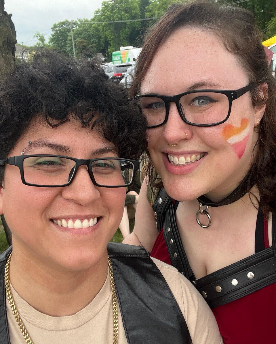 Rooster on the right smiling, wearing leather vest. Sol on the left smiling, wearing leather harness and lesbian pride color heart on cheek.