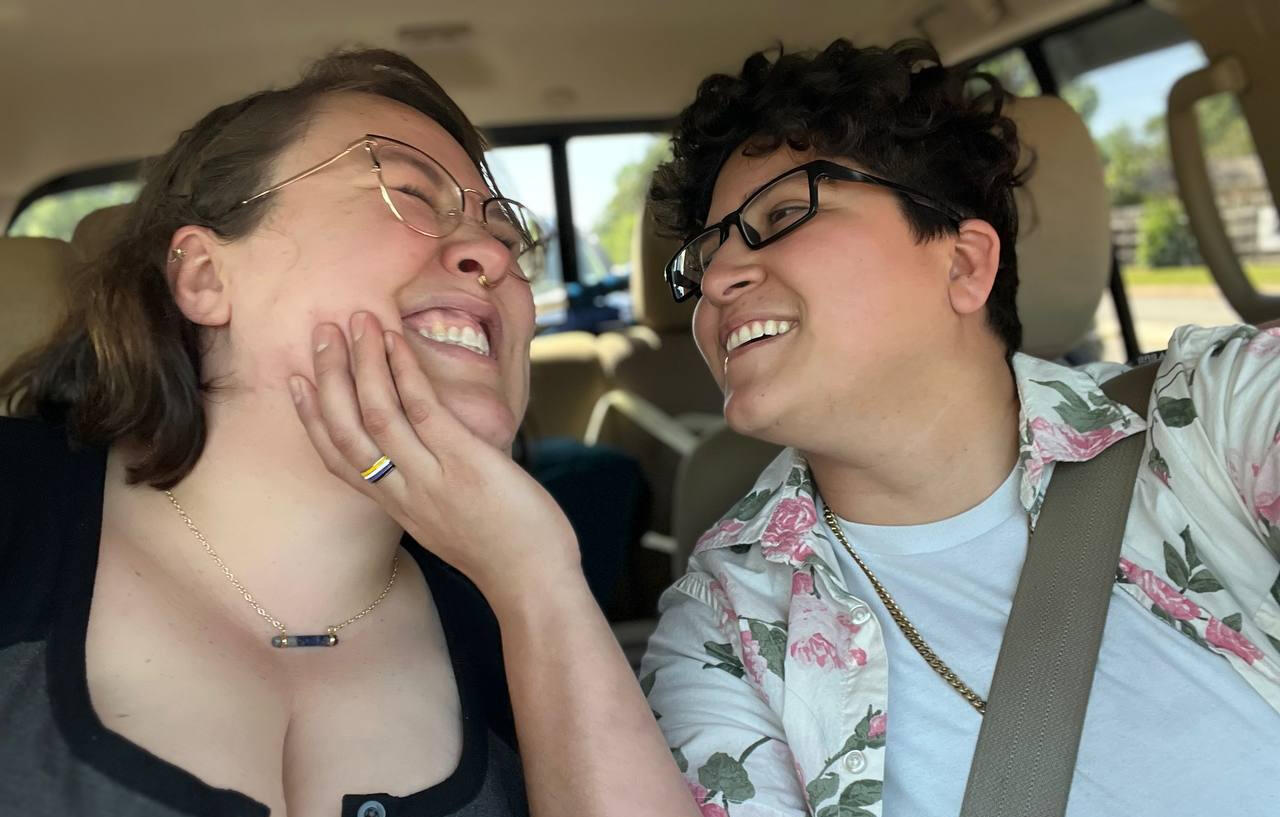 Sol on the left, smiling while their face is being held by Rooster. Rooster on the right, smiling while holding Sol's chin in one hand.